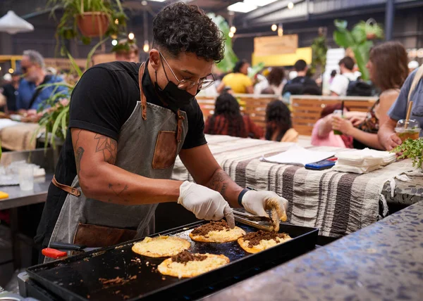 A multi-ethnic chef spoons meet onto tacos at a busy indoor food market. High quality photo