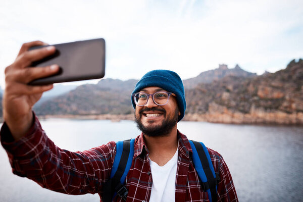 A happy hiker takes a selfie using smartphone with lake and mountains in the background. High quality photo