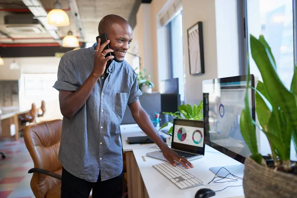 A Black man stands at his desk in the office answering a call while checking data. High quality photo