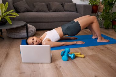 Young multi-ethnic woman does bridge exercise watching online fitness class. High quality photo