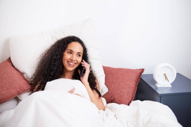 Genç, çok ırklı bir kadın, yataktan şehirlerarası samimi telefon konuşmalarında güler. Yüksek kalite fotoğraf