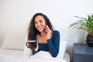 Genç, çok ırklı bir kadın yataktan telefonla konuşurken gülüyor. Yüksek kalite fotoğraf