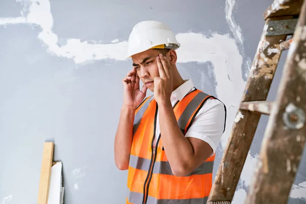 A young Asian construction worker rubs his temples in pain on site. High quality photo