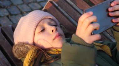 Parktaki bankta uzanan kız çocuk akıllı telefon aygıtı kullanıyor ve tembel bir hayatı var.