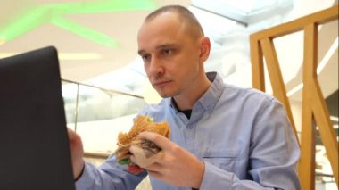 Şehirdeki bir alışveriş merkezinde çalışan genç adam büyük lezzetli hamburgerler yiyor ve dizüstü bilgisayarı çiğniyor.