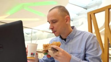 WROCLAW, POLAND - Şubat 03, 2022: Bir alışveriş merkezinde uzaktan çalışan genç adam büyük lezzetli hamburgerler yiyor, kola bardağı çiğniyor ve dizüstü bilgisayar dakları yazıyor.