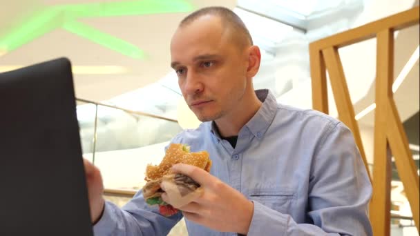 Mladý muž pracuje na dálku z městského obchoďáku potravin soud jíst velké chutné burger žvýkání a psaní notebook
