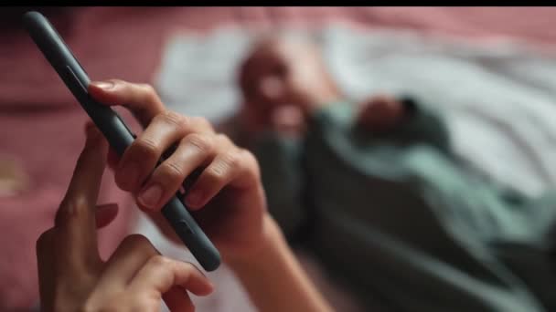 Moeder is aan de telefoon terwijl haar baby ligt op het bed en bewegende handen — Stockvideo
