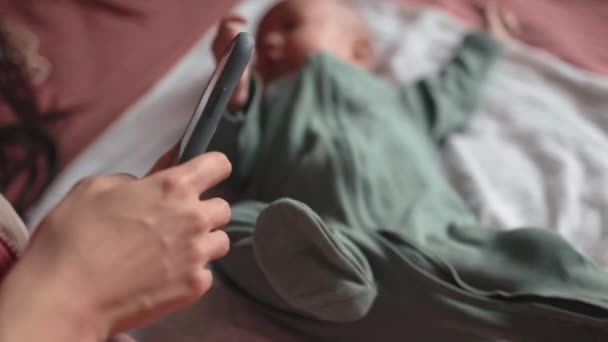 Мама разговаривает по телефону, пока ее ребенок лежит на кровати и двигает руками. — стоковое видео