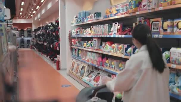 Asian woman in white choosing goods in supermarket with stroller — Vídeos de Stock