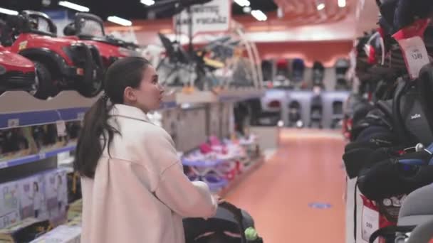 Asian woman in white choosing car seats in supermarket with stroller — Vídeos de Stock