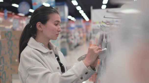 Asian woman in white choosing baby bibs in supermarket — Stockvideo