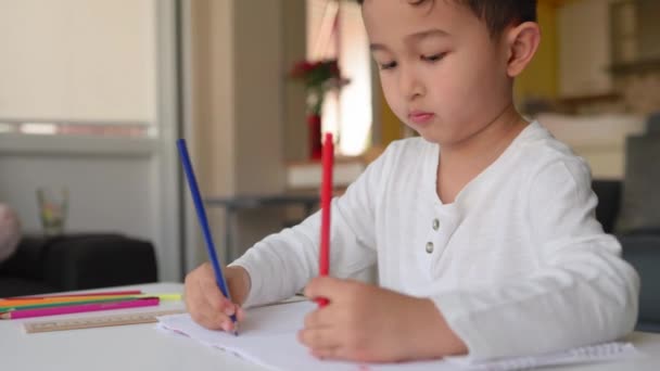 Маленький азіатський хлопчик на білому малюнку, використовуючи обидві руки з синьою і червоною ручкою на папері, сидячи вдома — стокове відео
