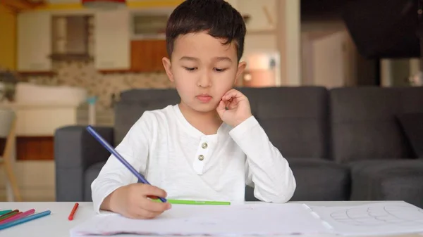 Mały azjatycki chłopiec w białym rysunku z niebieskim piórem na papierze siedzi w domu — Zdjęcie stockowe