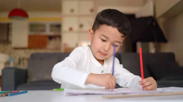 Little asian boy in white drawing using both of hands with blue and red pen on paper sitting at home — 스톡 사진