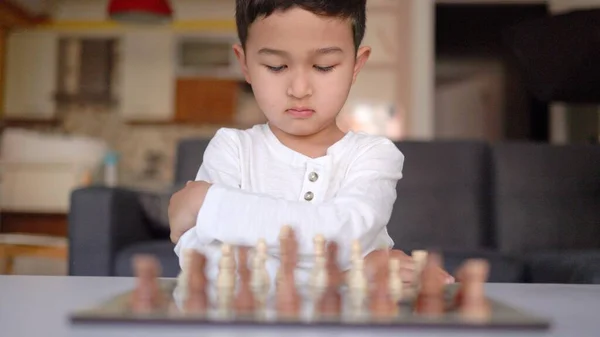 Μπροστά άποψη του μικρού αγοριού στα λευκά παίζει σκάκι στο τραπέζι — Φωτογραφία Αρχείου