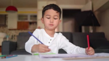 Little asian boy in white drawing using both of hands with blue and red pen on paper sitting at home