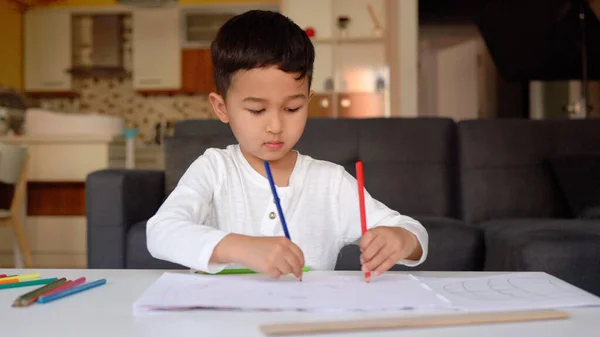 Little asian boy in white drawing using both of hands with blue and red pen on paper sitting at home — Fotografia de Stock