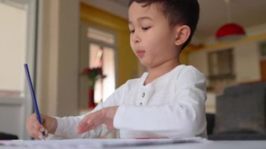 Beyaz çizim yapan küçük Asyalı çocuk. Elinde mavi kalemle evde oturuyor.