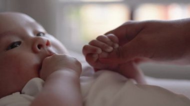 Cute baby in white holding mothers hand and sucks his thumb 