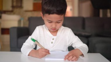 Beyaz yazılı küçük Asyalı çocuk. Elinde mavi kalemle evde oturuyor.