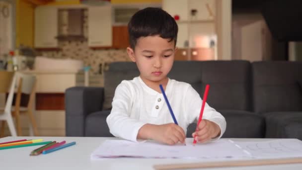 Pequeno menino asiático em desenho branco usando ambas as mãos com caneta azul e vermelha no papel sentado em casa — Vídeo de Stock