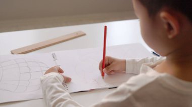 Beyaz kağıda kırmızı kalem çizen anaokulu öğrencisinin arka görünümü.