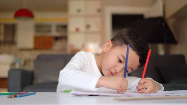 Little asian boy in white drawing using both of hands with blue and red pen on paper sitting at home — стоковое фото