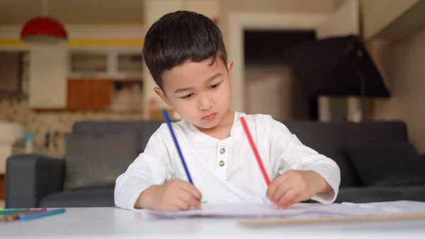 Little asian boy in white drawing using both of hands with blue and red pen on paper sitting at home — Photo