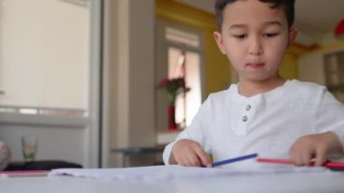 Evinde oturan mavi ve kırmızı kalemli beyaz çizim yapan küçük Asyalı çocuk.