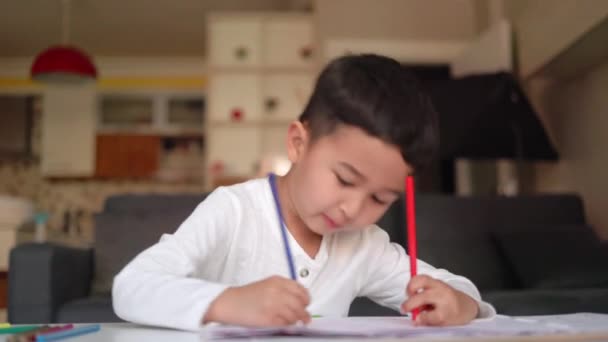 Beyaz çizim yapan küçük Asyalı çocuk, elinde mavi ve kırmızı kalemle evde oturuyor. — Stok video