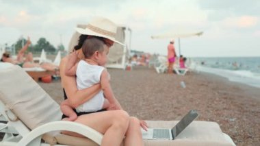 Freelancer woman with baby working on laptop near the sea. Remote work concept. work on vacation. 4 k. High quality 4k footage