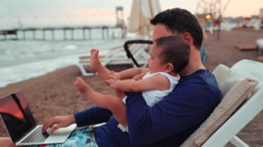 Freelancer man with baby working on laptop near the sea . Remote work concept. work on vacation. 4 k. High quality 4k footage