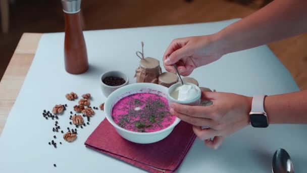 Mano poner crema agria en sopa de remolacha fría con pepino y eneldo en tazón blanco — Vídeos de Stock