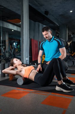 Genç bir bayan, spor salonunda antrenörle miyofasyal gevşeme tekerleğiyle rahatlıyor.