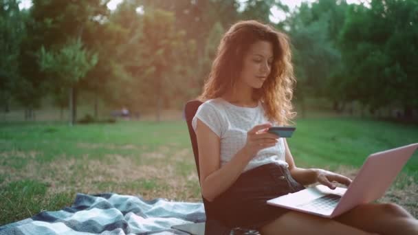 Jovem fêmea fazer compra online com cartão de crédito e laptop no parque ao ar livre — Vídeo de Stock