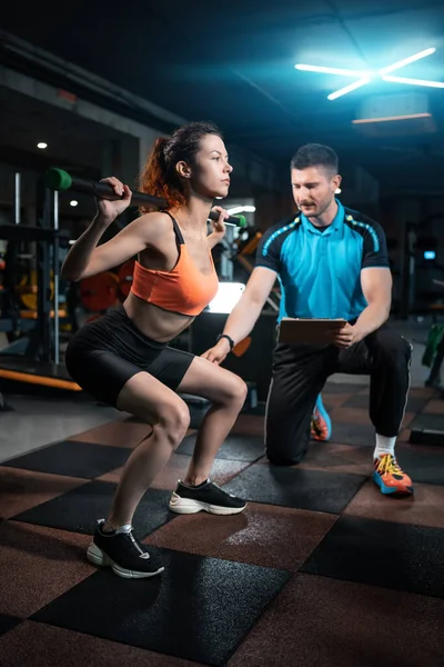 Jonge vrouwelijke training en het trainen van lange halter squat in de sportschool met personal trainer — Stockfoto