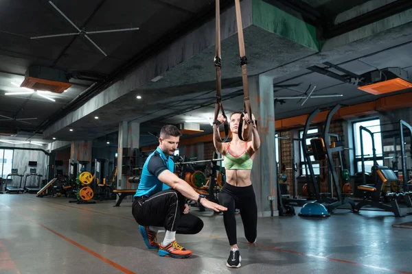 Genç bayan spor yapıyor ve spor salonunda Trx ve kişisel antrenörle antrenman yapıyor.