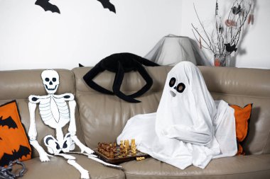 Hayalet kostümü giymiş bir çocuk kanepeye oturur ve bir iskeletle satranç oynar. Komik Cadılar Bayramı. Cadılar Bayramı kartpostalı. Mutlu bir çocukluk. Dekorasyon ve parti konsepti. Geleneksel sonbahar festivali. Modifiye et ve uzayı kopyala.