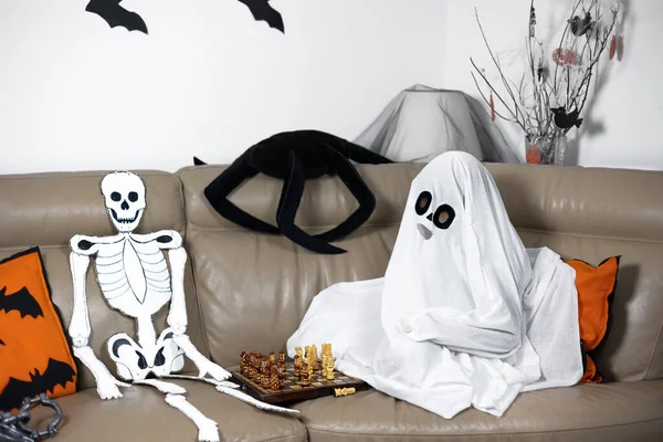 Hayalet kostümü giymiş bir çocuk kanepeye oturur ve bir iskeletle satranç oynar. Komik Cadılar Bayramı. Cadılar Bayramı kartpostalı. Mutlu bir çocukluk. Dekorasyon ve parti konsepti. Geleneksel sonbahar festivali. Modifiye et ve uzayı kopyala.