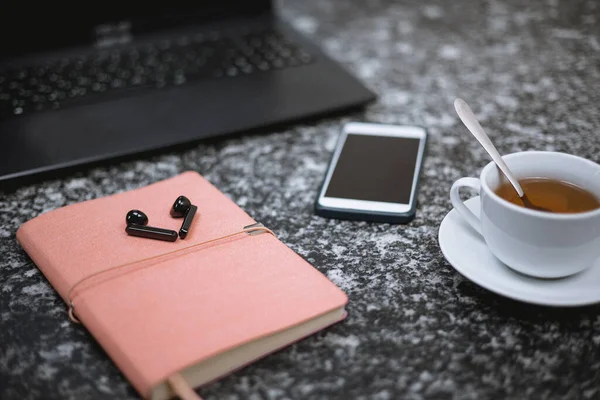 On the black marble table are a laptop, wireless headphones a cell phone, a notepad, a cup of tea.Selective focus business man desktop. On the table are cell phone, wireless headphones a notepad.