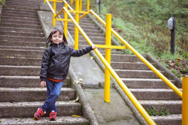 Pilot şapkalı ve kahverengi ceketli mutlu çocuk bir sonbahar parkında merdivenlerde duruyor ve hayaller kuruyor. Çocuk modası.