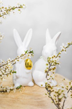 Pençelerinde altın yumurtayla çiçek açan kiraz dallarındaki ahşap palette iki beyaz seramik tavşan. Paskalya konsepti.