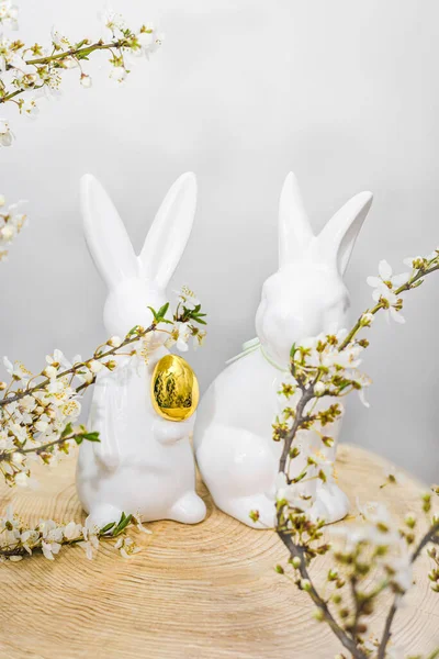 Two white ceramic rabbits on a wooden pallet in blossoming cherry branches with a golden egg in their paws. Easter concept.