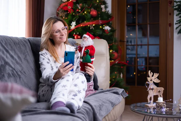 Young positive pretty woman in knitted winter warm socks and pajama sitting on couch and talking on the phone. New Year and Christmas decoration. Online distant remote communication.