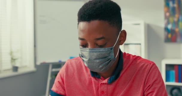 顔に保護マスクを持つ暗い肌の若い男の子は野心的なプロジェクトに取り組んでいます,コンピュータの前に座って焦点を当て,ポンダー,側面に見えます,最先端の画面に視線を返します,. — ストック動画