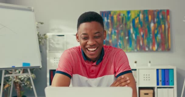 Um jovem menino de beleza afro-americana senta-se à mesa em uma sala de escritório com um laptop, sorri amplamente e acena a mão, continua uma conversa via internet, vídeo, tem ensino remoto. — Vídeo de Stock