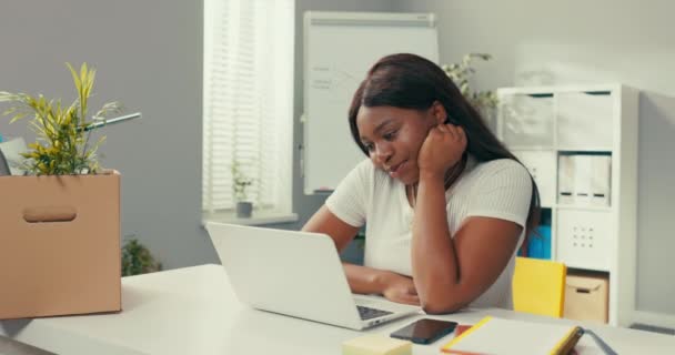 Bescheidenes, hübsches, dunkelhäutiges Mädchen sitzt im Zimmer am Schreibtisch, verbringt den Nachmittag mit Freunden, surft im Internet, neben dem Telefon, Notebooks, Frauenrequisiten unterm Kinn, lächelt sanft auf den Laptop-Bildschirm. — Stockvideo