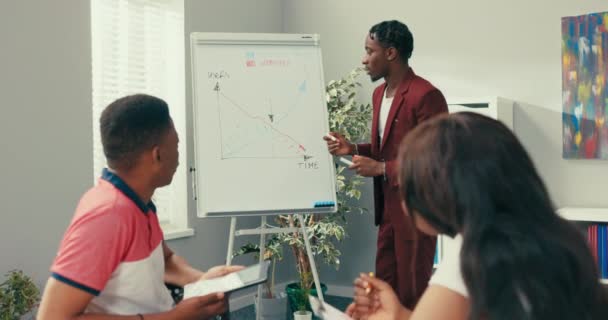 Un employé expérimenté de longue date d'une grande entreprise se tient au bureau par tableau blanc dessine un tableau mène une formation pour les stagiaires, les jeunes futurs employés prennent des notes tout en écoutant des hommes à la peau foncée — Video