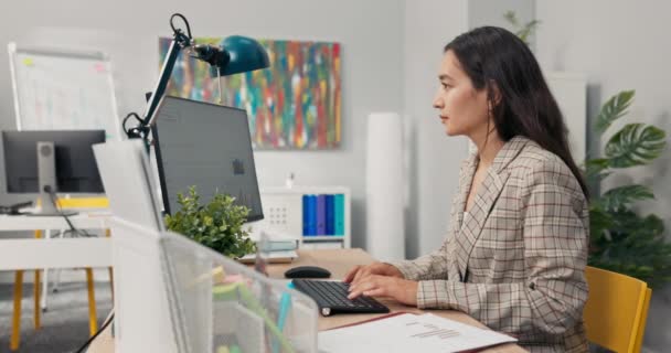 An attractive, experienced company accountant sits in office in front of a computer checking statistical data employee salary charts comparing paper diagrams with electronic ones financial analyst. — Stock Video